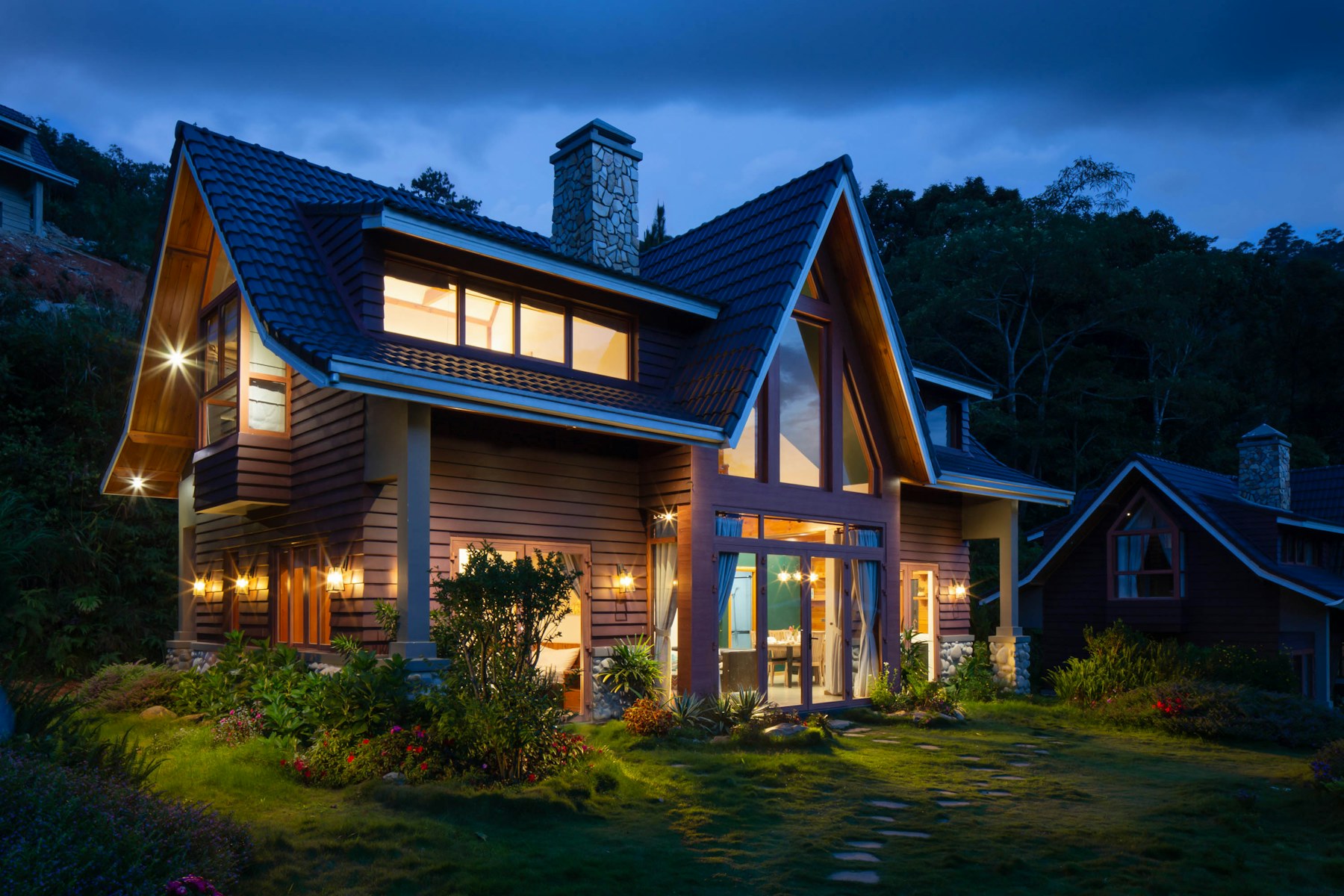 An illuminated two-story home with a tiled roof and landscaped garden at dusk